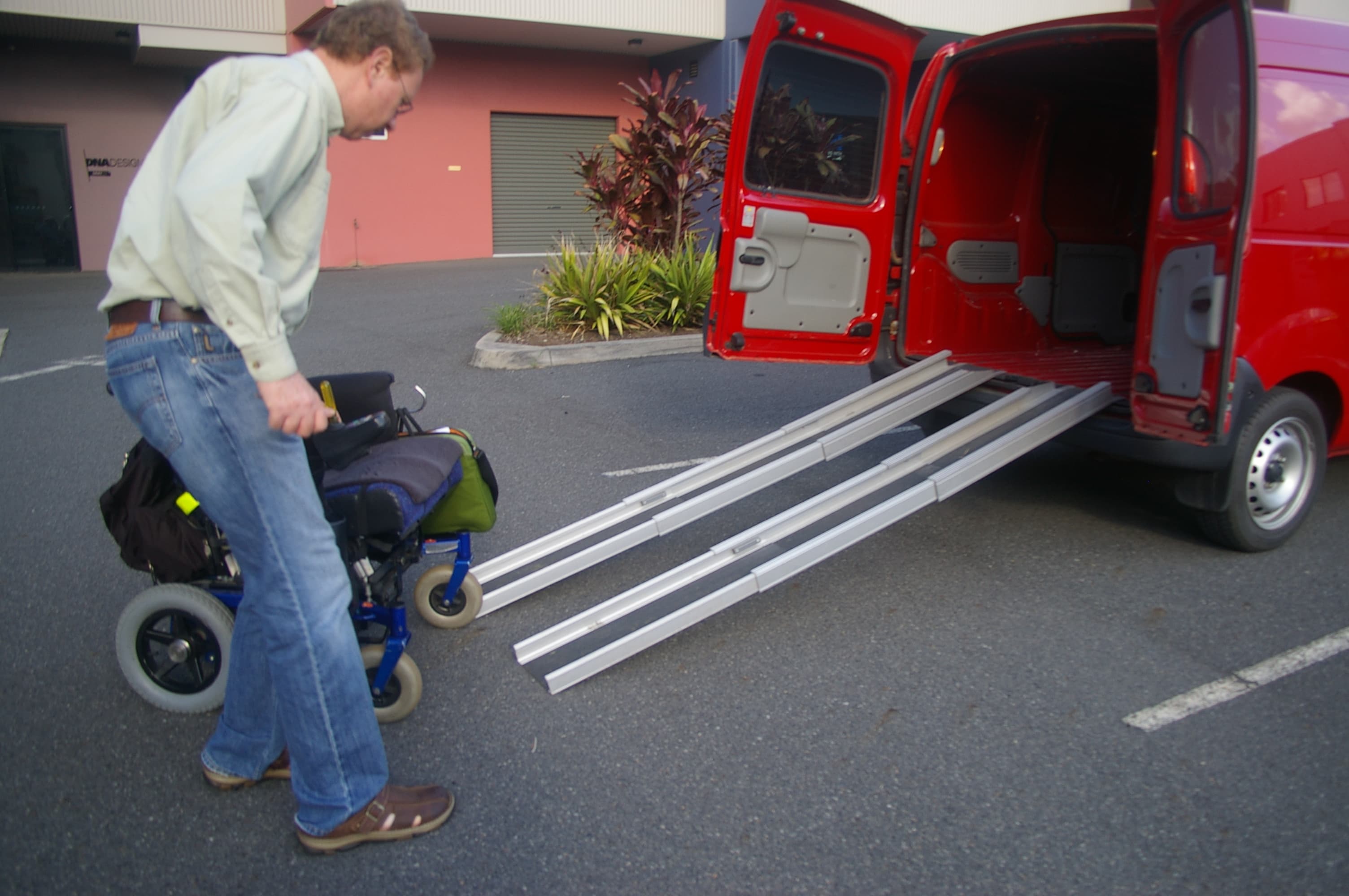 A powered wheelchair being guided up a rear loading ramp into a vehicle in a paved parking area
